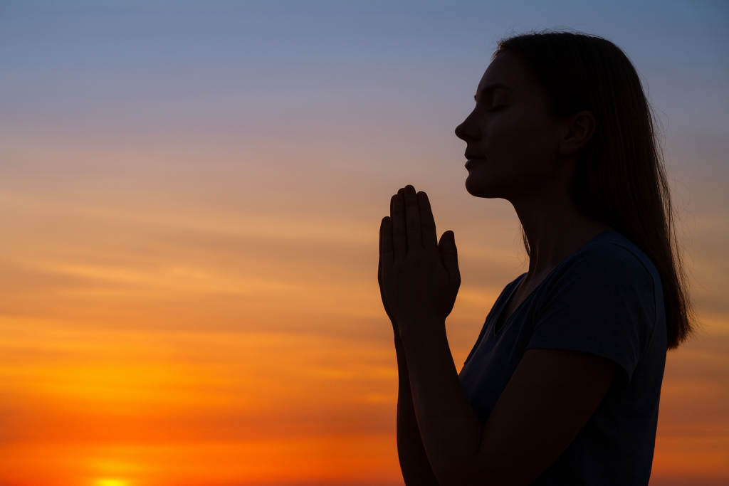 A woman praying with hands clasped at sunset, silhouetted against a colorful sky, symbolizing daily prayer and talking to God like a friend.